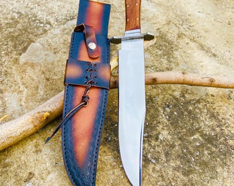 May include: A brown leather sheath with a decorative stitch pattern and a silver button. A silver and steel knife with a wooden handle is in the sheath. The knife and sheath are on a brown and gray textured surface.