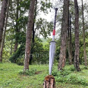 May include: A large, silver sword with a red and black handle is displayed outdoors, resting on a tree stump. The sword's blade is wide and flat, and the handle has a textured grip. The background features a forest with tall trees and green foliage.