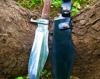 May include: A large, polished steel knife with a wooden handle and a black leather sheath. The knife blade is highly reflective, and the handle is a dark brown color. The sheath has gold-colored button closures. The knife is displayed against a natural background.