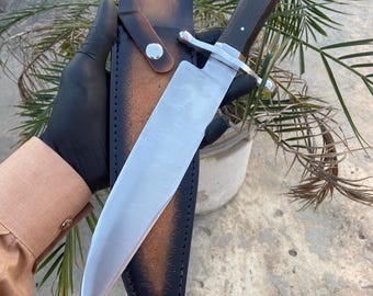 May include: A silver Bowie knife with a black wooden handle and a leather sheath. The knife is held in a gloved hand.