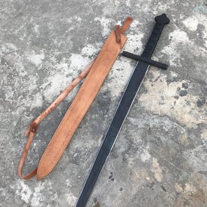 Può includere: Una spada nera con guardia e fodero in pelle. La spada ha una lama lunga e dritta e un'impugnatura nera. Il fodero è marrone e ha un cinturino per il trasporto. La spada è lunga circa 91 cm.
