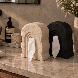 May include: Two arched tissue holders, one in a textured beige stone finish and one in a textured black stone finish, each featuring a central opening with a white tissue protruding from the centre, displayed side by side on a marble countertop.