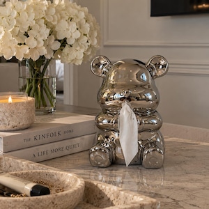 May include: A shiny, silver-coloured, bear-shaped tissue dispenser. The bear is depicted in a seated position with its hands held together near its face, where a single white tissue is pulled through a small opening in the centre of its mouth.