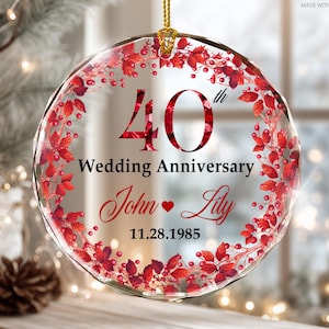 May include: A clear, round glass ornament with a gold hanging loop. The ornament features the text "40th Wedding Anniversary" and the names "John & Lily" with a date. Red floral accents and a decorative border complete the design.