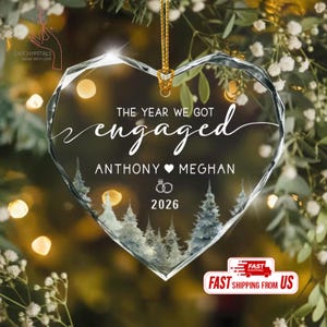 May include: A heart-shaped clear glass ornament featuring the text THE YEAR WE GOT engaged, the names ANTHONY MEGHAN, a ring icon, and the year 2026. The lower portion displays a printed graphic of evergreen trees, suspended by a gold cord.