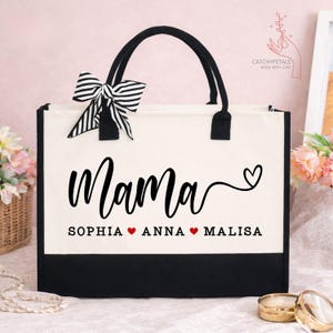 May include: A canvas tote bag with black handles and a black bottom. The word "Mama" is written in a cursive font, with the names Sophia, Anna, and Malisa below. A black and white striped bow is attached.