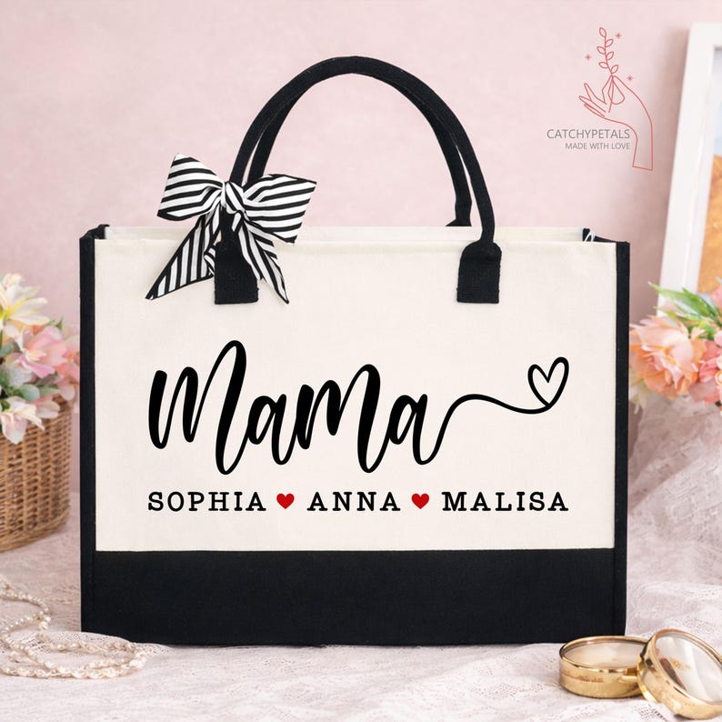 May include: A canvas tote bag with black handles and a black bottom. The word "Mama" is written in a cursive font, with the names Sophia, Anna, and Malisa below. A black and white striped bow is attached.