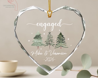 May include: A clear, heart-shaped ornament with the word "engaged" in white script. Below are three watercolor Christmas trees and the names "Mike & Veronica" with the year "-2026". A gold chain is attached for hanging.