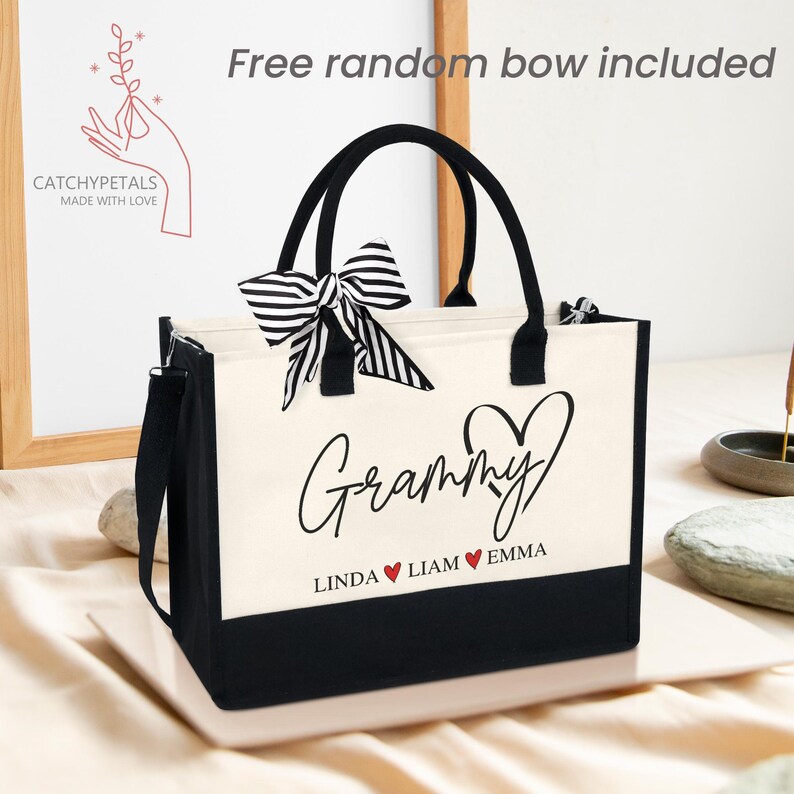 May include: A canvas tote bag with black handles and a black bottom panel. The bag features the word "Grammy" in script, with the names "Linda", "Liam", and "Emma" below. A black and white striped bow is attached to the handle.