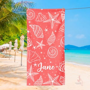 Pu&ograve; includere: Asciugamano da spiaggia rosa corallo con un motivo di conchiglie e stelle marine bianche. L'asciugamano ha il nome "Jane" scritto in bianco.