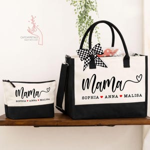 May include: A canvas tote bag and matching cosmetic bag set. Both bags are white with black bottoms and feature the word "Mama" in script, along with names. The tote has black handles and a checkered bow.