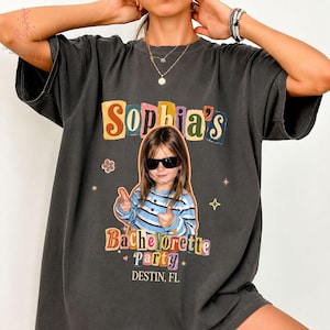 May include: A charcoal gray short-sleeve t-shirt featuring a graphic print of a child wearing sunglasses and a blue striped shirt. The text reads Sophia's Bachelorette Party DESTIN, FL, surrounded by colorful, retro-style block letters and stars.