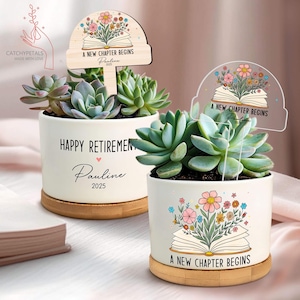 May include: Two white ceramic succulent planters with wooden bases. One planter reads "Happy Retirement Pauline 2025" and the other reads "A New Chapter Begins Pauline 2025". Both planters feature a succulent plant and a decorative sign.