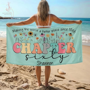 May include: A turquoise beach towel with the text "Making the world a better place since 1966" and "CHAPTER sixty Shannon" in colorful retro lettering. The towel features a floral design and is held by a person on a sandy beach.