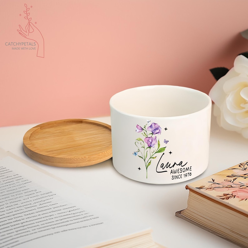May include: A white ceramic planter with a floral design featuring purple flowers, green leaves, and the name "Laura" with "Awesome Since 1976." A round wooden saucer sits beside the planter. The Catchy Petals logo is in the upper left corner.