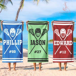 May include: Three personalized lacrosse towels in blue, green, and red. Each towel features a lacrosse helmet and crossed sticks design, a name, and a number. The towels are held up on a beach with a blue sky and palm trees in the background.