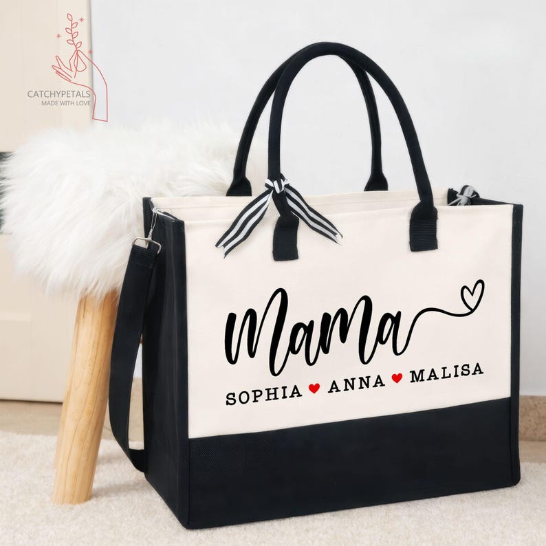 May include: A canvas tote bag with black handles and a black bottom panel. The bag features the word "Mama" in cursive, with the names Sophia, Anna, and Malisa below. A heart symbol is between each name. The bag has a detachable shoulder strap.