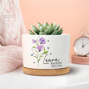 May include: A white ceramic planter with a succulent plant, a bamboo base, and a floral design with the name "Laura" and the text "AWESOME SINCE 1976". The planter has a round shape.