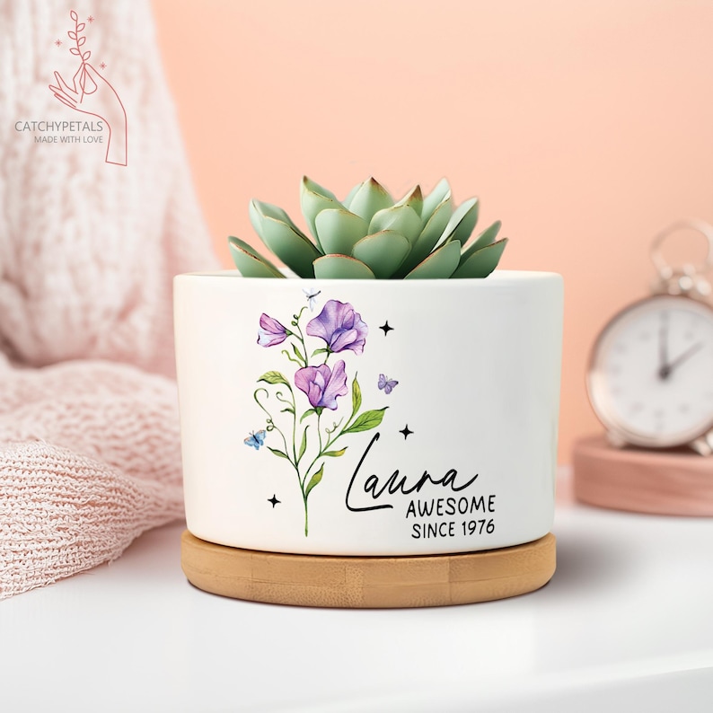 May include: A white ceramic planter with a succulent plant, a bamboo base, and a floral design with the name "Laura" and the text "AWESOME SINCE 1976". The planter has a round shape.