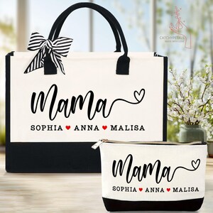 May include: A canvas tote bag and matching cosmetic bag set. The bags are off-white with black accents and handles. The word "Mama" is printed in black script, along with three names and hearts. A black and white striped bow adorns the tote bag.