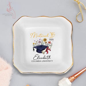 May include: White ceramic trinket dish with a gold rim. It features a graduation cap with a diploma, flowers, and the text "Mastered It 2025" and "Elizabeth COLUMBIA UNIVERSITY".