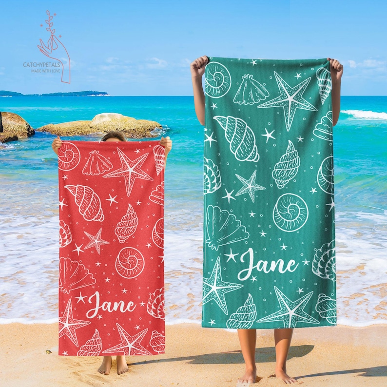 Pu&ograve; includere: Due asciugamani da spiaggia con un motivo a conchiglie e stelle marine. Gli asciugamani sono rossi e turchesi con contorni bianchi. Il testo "Jane" &egrave; scritto su ogni asciugamano.