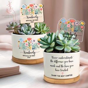 May include: Two white ceramic succulent pots with floral designs and wooden bases. Each pot features a succulent plant and a personalized sign that reads "Kimberly Officially Retired 2025." One pot has a message: "Never underestimate the difference you have made and the lives you have touched."