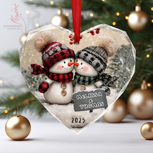 May include: Heart-shaped Christmas ornament with two snowmen in hats and scarves. The snowmen are kissing, with a sign reading "Melissa & Thomas" and the year "2025". The ornament is suspended by a red ribbon.
