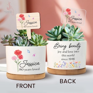 May include: Two white ceramic succulent planters with wooden bases. The front planter has a floral design with the name "Jessica" and "80 years loved." The back planter says "Bring beauty" and "joy and love into this world since 1946."