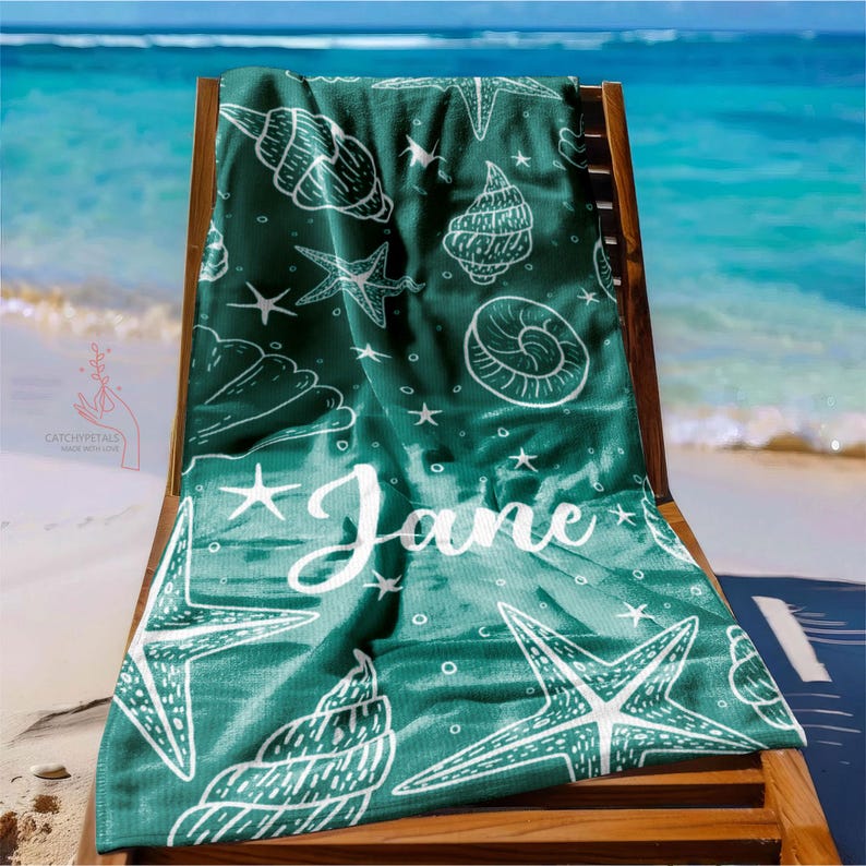 Pu&ograve; includere: Un asciugamano da spiaggia color turchese con un motivo di conchiglie e stelle marine bianche. L'asciugamano ha il nome "Jane" scritto in lettere bianche corsive.