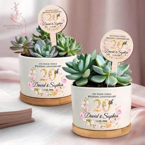 May include: Two white ceramic pots with succulents, each featuring a wooden sign and a bamboo saucer. The pots are decorated with floral designs and text that reads "On Your China Wedding Anniversary" and "20th". The names "David & Sophia" and the date "12.08.2006" are also displayed.