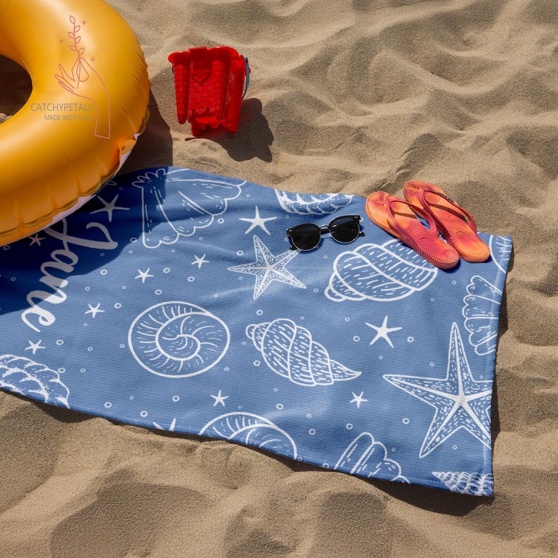 Pu&ograve; includere: Un asciugamano da spiaggia blu con un motivo a conchiglie e stelle marine bianche. L'asciugamano ha la parola "Have" scritta in bianco. L'asciugamano &egrave; sdraiato su una spiaggia sabbiosa con un anello gonfiabile giallo, un secchiello rosso e infradito.