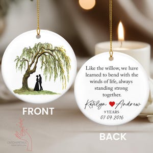 May include: A white, round Christmas ornament with a gold string. The front features a watercolor painting of a weeping willow tree and a couple. The back has a quote and names: Katelyn & Andrew, 9 years, 07.09.2016.