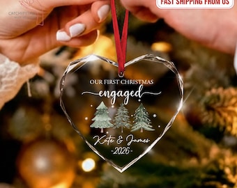May include: A heart-shaped clear acrylic ornament featuring a red ribbon hanger. The text reads OUR FIRST CHRISTMAS engaged, followed by the names Kate & James and the year 2026. Three illustrated evergreen trees are printed below the text.