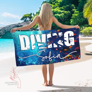 May include: A rectangular beach towel with the word "DIVING" in large white block letters, a diver silhouette, and the name "Sofia" in cursive. The towel features an underwater scene with fish and coral, set against a blue background.