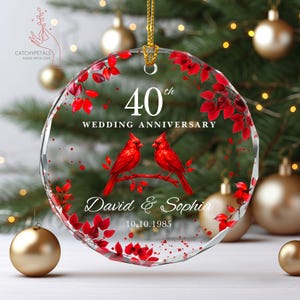 May include: A clear, round ornament with a gold hanging loop. The ornament features red cardinals perched on a branch, red leaves, and the text "40th Wedding Anniversary" with names and a date. Gold ornaments and a Christmas tree are in the background.