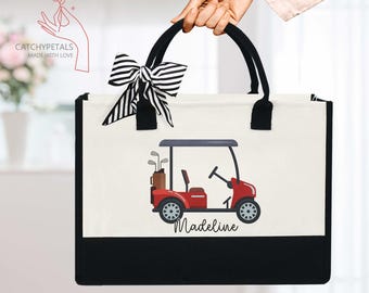 May include: A white canvas tote bag with black handles and a black bottom. The bag features a red golf cart with the name "Madeline" printed below it. A black and white striped bow is tied to the handles.