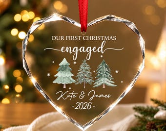 Może przedstawiać: Ozdoba w kształcie serca z przezroczystego szkła o fasetowanych krawędziach, zawieszona na czerwonej wstążce. Zawiera napis "OUR FIRST CHRISTMAS engaged", imiona "Kate & James" oraz rok "2026" nad trzema choinkami w stylu akwareli.