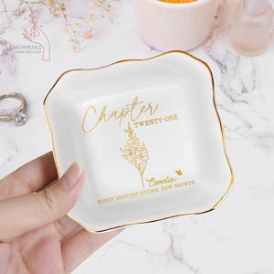 May include: A white ceramic trinket dish with gold trim, held by a hand. The dish features the words "Chapter Twenty-One" and "Camellia" with a floral illustration in gold. The text "Every Chapter Brings New Growth" is also present.