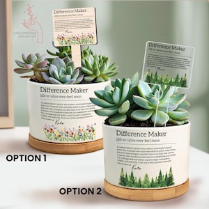 May include: Two white ceramic succulent planters with wooden bases, each featuring the text "Difference Maker" and a definition. One planter has a floral design, the other a forest design. Both have a wooden sign.