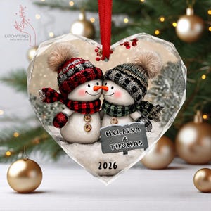 May include: A heart-shaped Christmas ornament featuring two snowmen wearing knit hats and scarves. One snowman wears red plaid, the other green plaid. A gray sign reads MELISSA & THOMAS, with the year 2026 printed below. It hangs from a red ribbon.