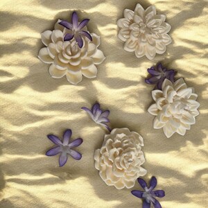 May include: Five white ceramic flowers with purple centers arranged on a textured beige surface. The flowers are in various sizes and shapes.