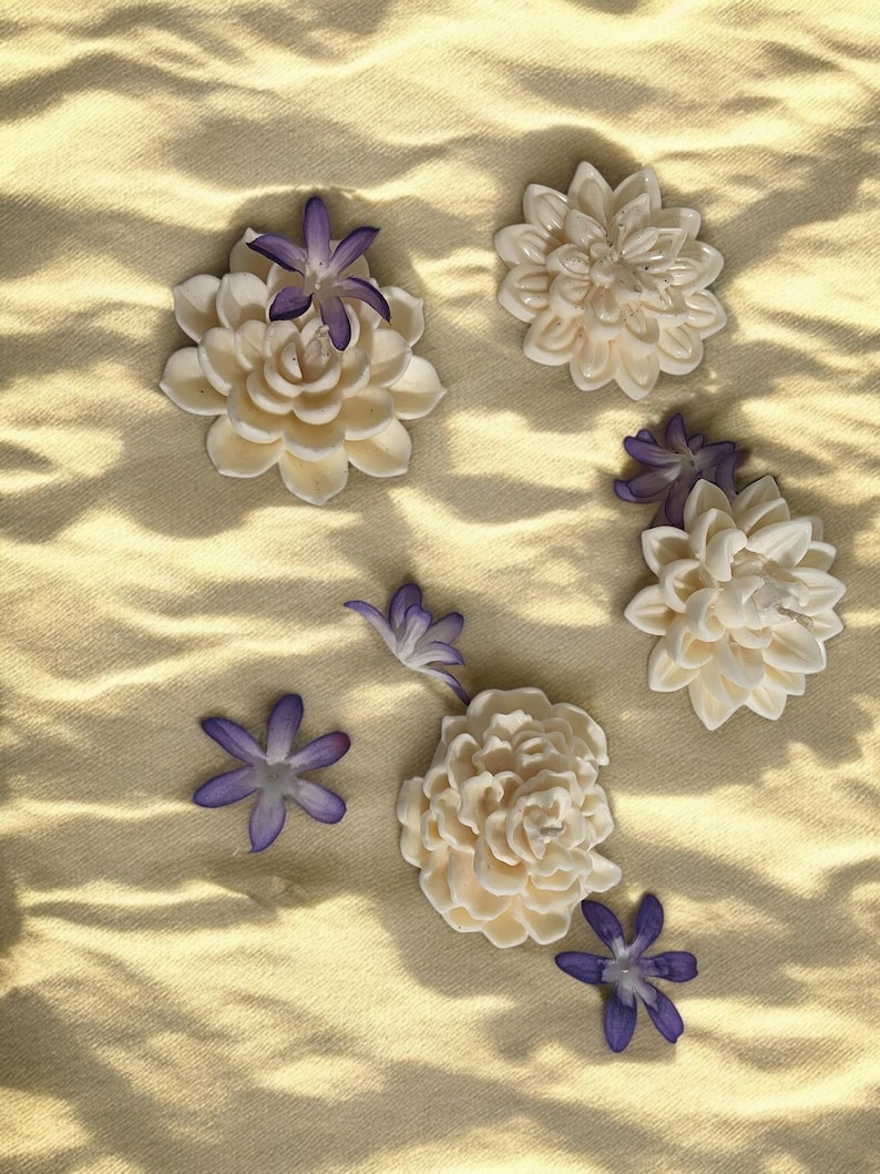May include: Five white ceramic flowers with purple centers arranged on a textured beige surface. The flowers are in various sizes and shapes.