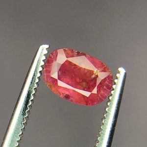 May include: A faceted, oval-shaped, red gemstone held by a pair of silver tweezers. The gem has a mix of red and orange hues, with visible internal inclusions. The background is a solid, neutral gray.