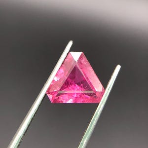 Puede incluir: Una gema triangular facetada de color rosa vibrante es sostenida por unas pinzas plateadas. El corte de la gema revela múltiples facetas, que capturan la luz y crean un efecto brillante. El fondo es un degradado de gris.