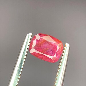 May include: A vibrant, cushion-cut ruby gemstone, held by silver tweezers. The ruby displays a rich, saturated red hue with internal light reflections. The gemstone's facets catch the light, showcasing its clarity and brilliance. The background is a neutral gray.