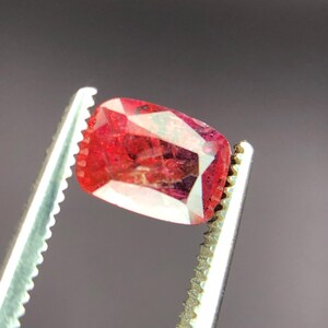 May include: A rectangular, faceted gemstone in a vivid red colour, held by silver tweezers. The gem exhibits internal patterns and a polished surface, demonstrating its clarity and brilliance. The tweezers provide a secure grip, highlighting the gem's shape and colour.