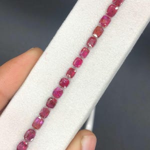 May include: A row of vibrant red gemstones, likely rubies, are displayed on a white surface. The gems vary slightly in shape, with a mix of square and oval cuts. The stones are held by a hand, showcasing their color and clarity.
