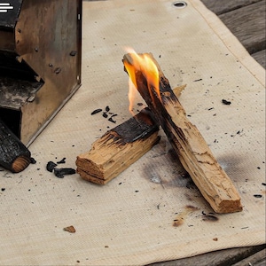 Può includere: Primo piano di ceppi di legno che bruciano con fiamme. I ceppi sono marrone chiaro con sezioni scure carbonizzate. Le fiamme sono arancioni e gialle. I ceppi sono su una superficie beige.