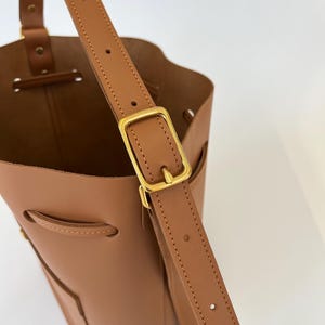 K&ouml;nnte beinhalten: Eine braune Ledereimer-Tasche mit einer goldenen Schnalle und einem verstellbaren Trageriemen. Die Tasche hat einen Kordelverschluss und eine kleine Tasche auf der Vorderseite.